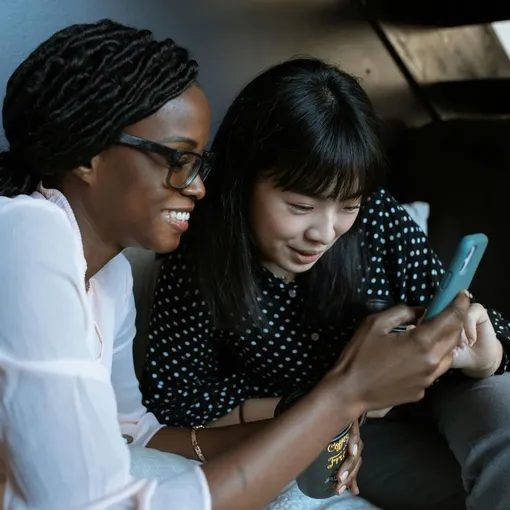 Two women smiling and engaging with social media content on a smartphone, representing connection and community online